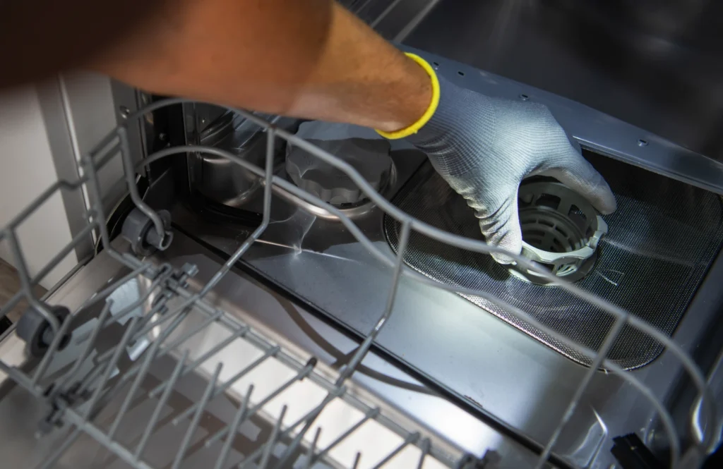 A gloved hand removing a dishwasher filter basket to clear hard water scale in a Midland, TX home kitchen.